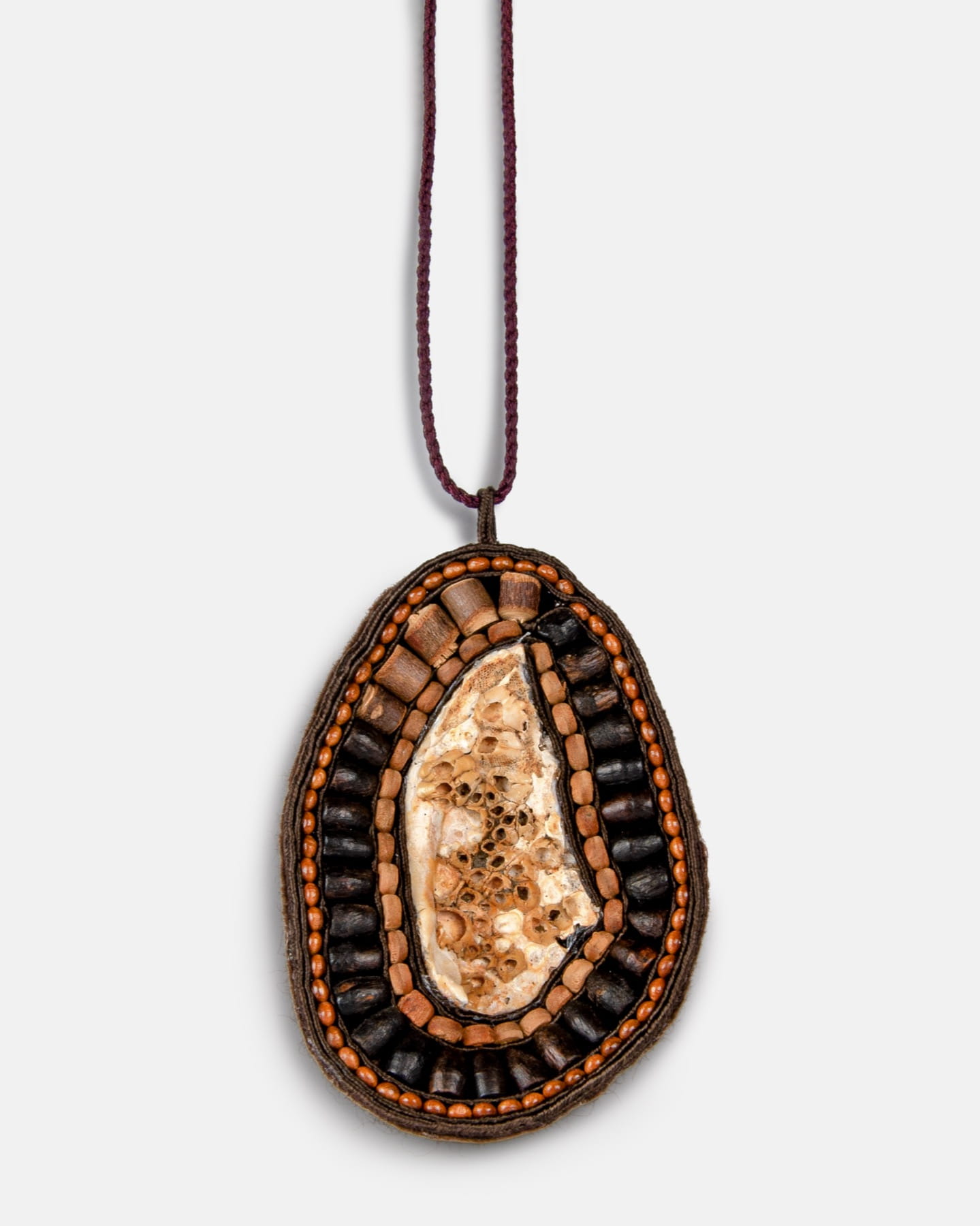 Amulet-wisiorek z naturalną muszlą morską ze Szkocji – ręcznie wykonany BG 16-3 Ręcznie wykonany wisiorek z centralnym beżowym kamieniem otoczonym warstwami brązowych, czarnych i pomarańczowych koralików, zawieszony na cienkim ciemnym sznurku na jednolitym jasnym tle.