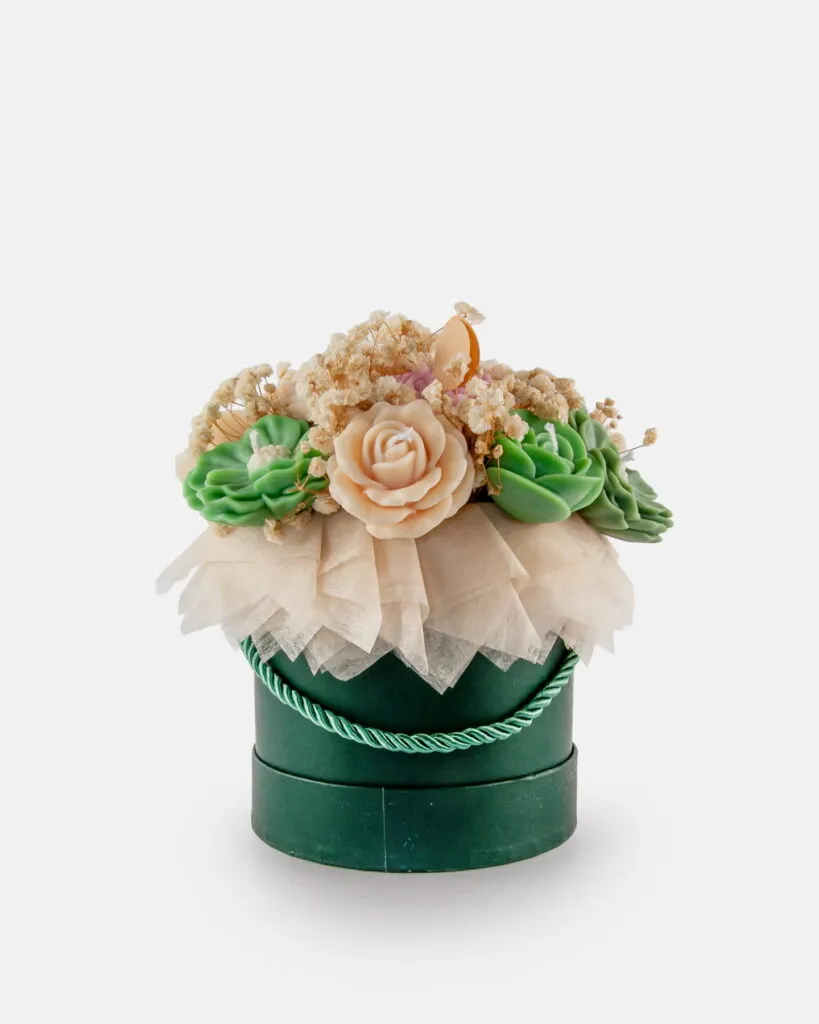 W zielonym, okrągłym pudełku z uchwytem znajduje się kompozycja ze sztucznych sukulentów, brzoskwiniowych kwiatów i beżowych suszonych kwiatów, owinięta warstwami beżowego papieru.