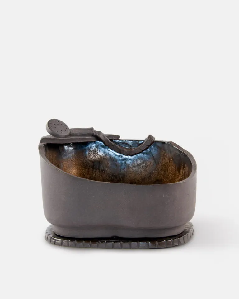 Ręcznie wykonana, owalna ceramiczna miseczka pokryta ciemnobrązową glazurą, z małą, dopasowaną łyżeczką opartą o jej brzeg. Miseczka stoi na teksturowanej, okrągłej podstawce.