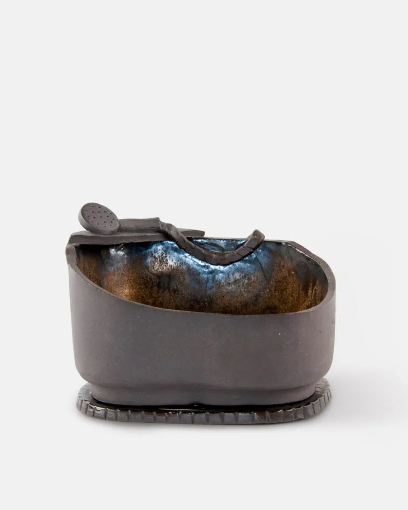 Ręcznie wykonana, owalna ceramiczna miseczka pokryta ciemnobrązową glazurą, z małą, dopasowaną łyżeczką opartą o jej brzeg. Miseczka stoi na teksturowanej, okrągłej podstawce.