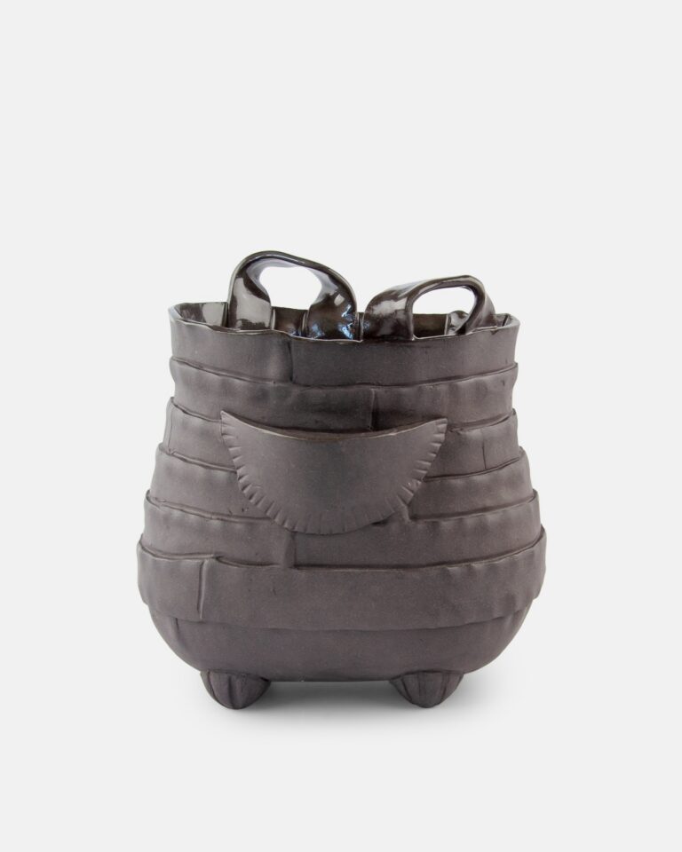 Czarna, rzeźbiarska ceramiczna waza o żebrowanej, zaokrąglonej bryle, oparta na małych nóżkach, z dwoma uchwytami u góry i ozdobnym wypukłym elementem z przodu.