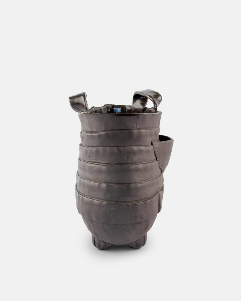 Ciemne, ręcznie wykonane naczynie ceramiczne o zaokrąglonym, żebrowanym korpusie, z trzema małymi nóżkami, bocznym uchwytem i dwoma nieregularnymi pętelkami na górze, na prostym białym tle.