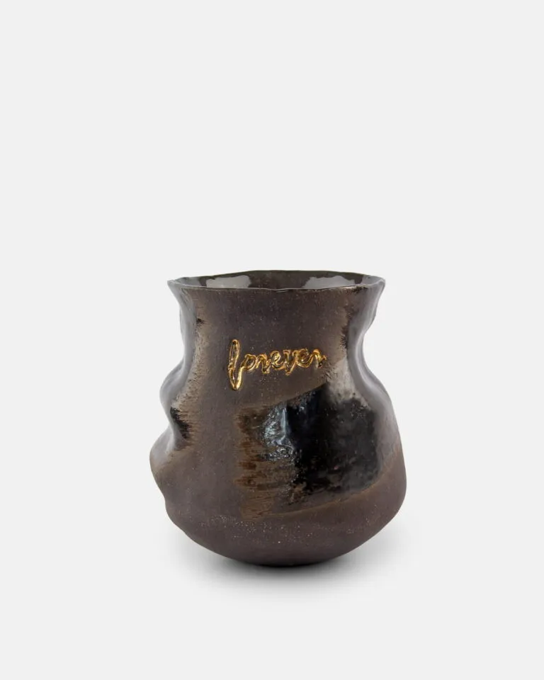 Błyszczący, ciemnobrązowy ceramiczny wazon o nieregularnych, organicznych kształtach, z napisem „forever” wykonanym złotą farbą na boku, na prostym białym tle.