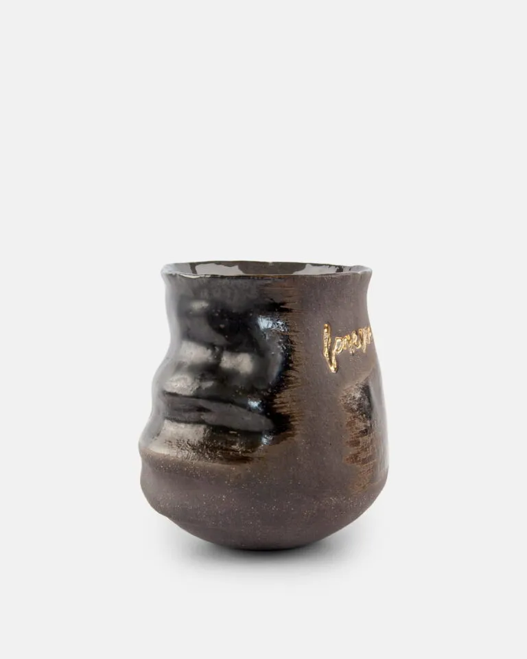 Mały, ciemny, błyszczący ceramiczny kubek o nierównej, falistej powierzchni, z napisem „love” wygrawerowanym złotem na jednej stronie. Tło jest gładkie i białe.