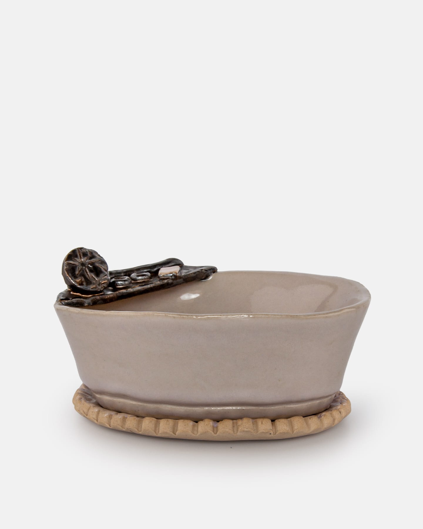 Ręcznie wykonana ceramiczna mydelniczka „Wanienka” KS 1-1 Beżowa ceramiczna miska z teksturowaną, okrągłą podstawą oraz dekoracyjnym, ciemnobrązowym akcentem przypominającym koło i pręty na jej krawędzi, na gładkim białym tle.