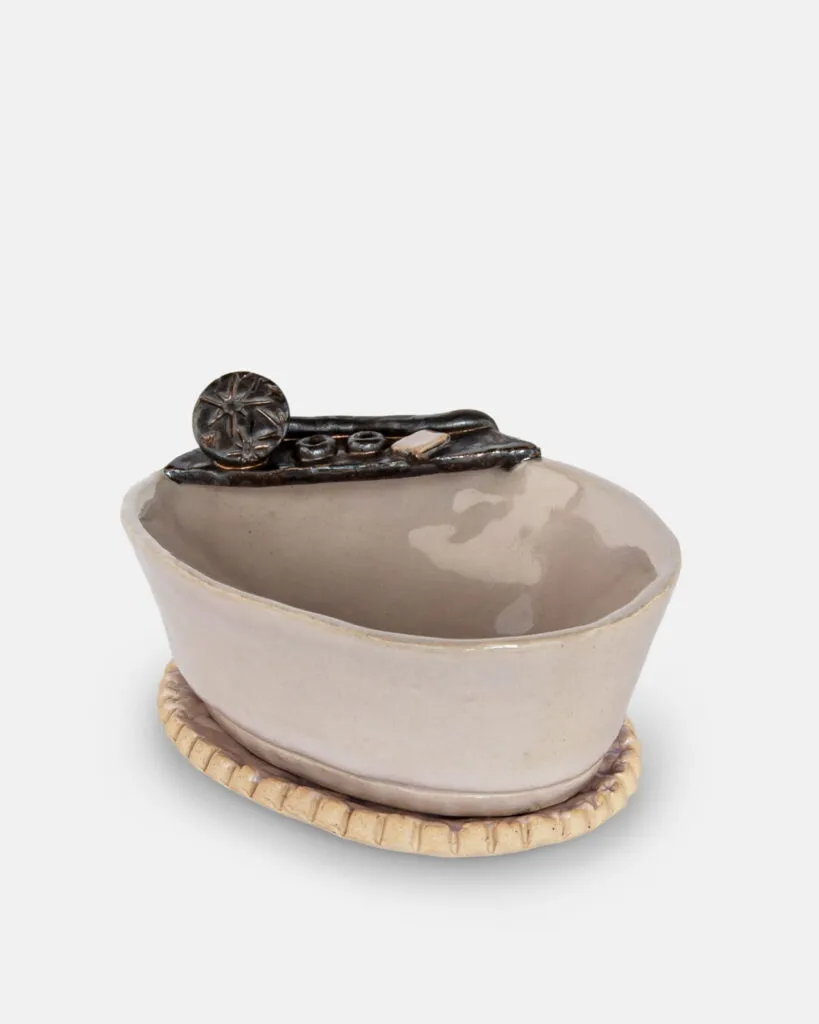 Ręcznie wykonana ceramiczna miska o nieregularnym owalnym kształcie, pokryta beżową glazurą i ozdobiona czarnym akcentem na krawędzi, stojąca na teksturowanej, okrągłej beżowej podstawie.