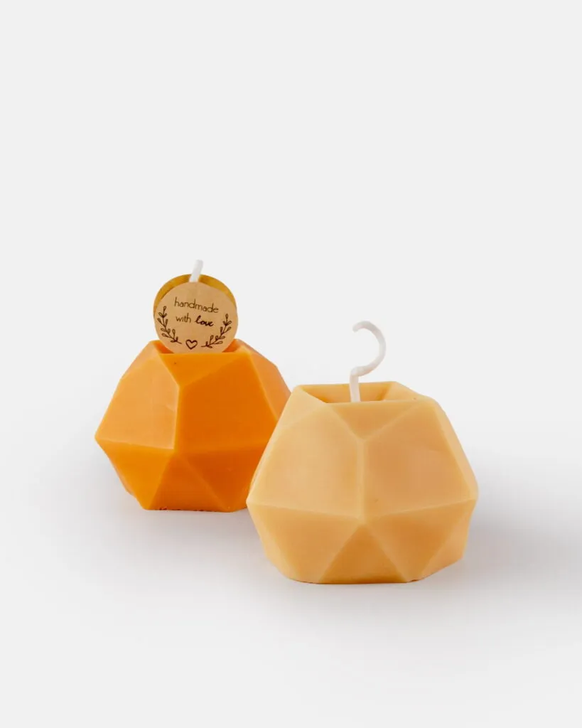 Dwie geometryczne, fasetowane świece w odcieniach pomarańczowym i jasnobeżowym. Jedna z nich ma okrągłą zawieszkę z napisem „handmade with love” oraz prosty knot; druga ma zakrzywiony knot. Obie świece są umieszczone na jednolitym białym tle.