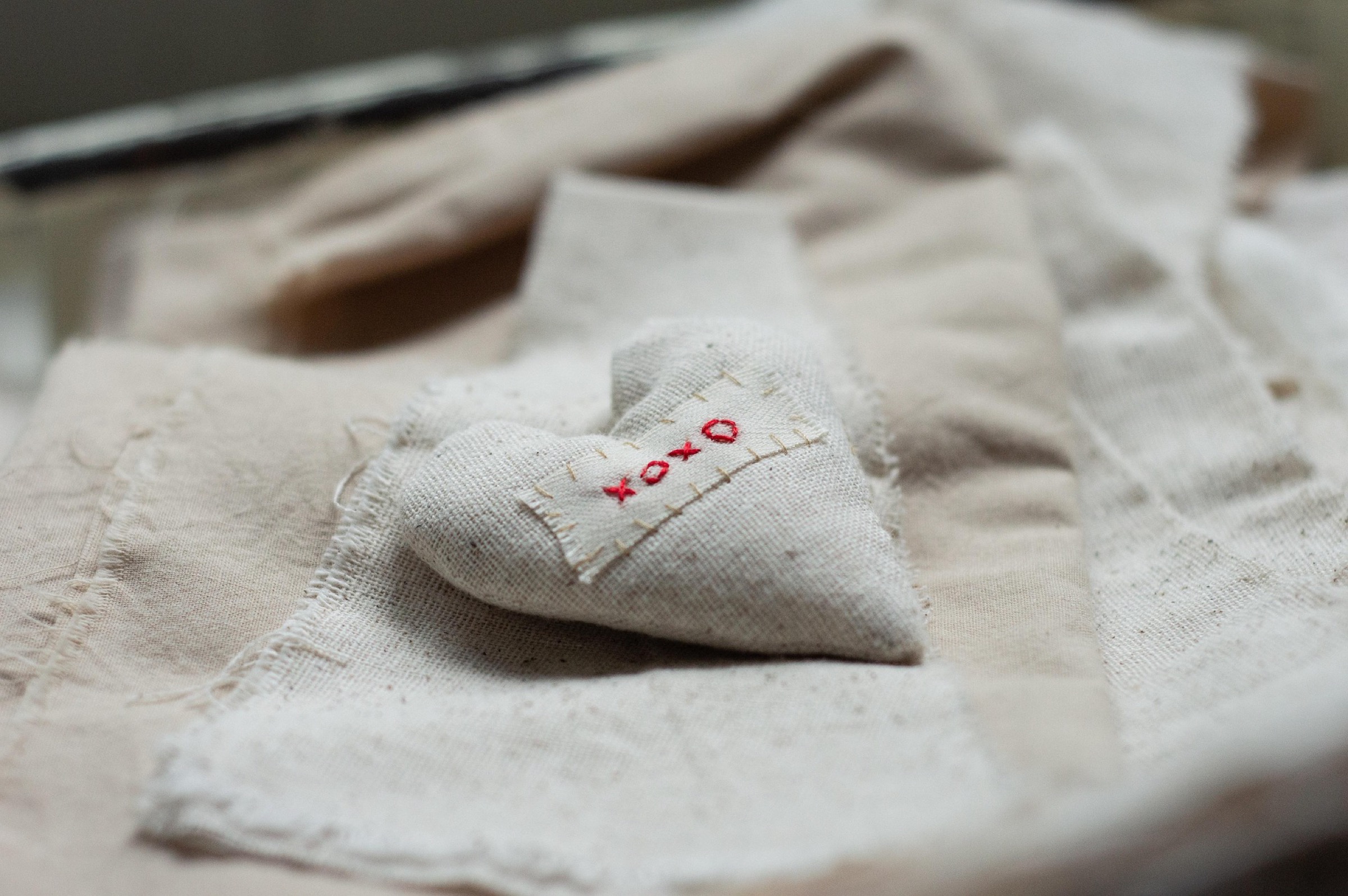 Małe, materiałowe serduszko z napisem „xoxo” wyszytym czerwoną nicią spoczywa na warstwach beżowej tkaniny, nadając mu delikatny, ręcznie wykonany wygląd.