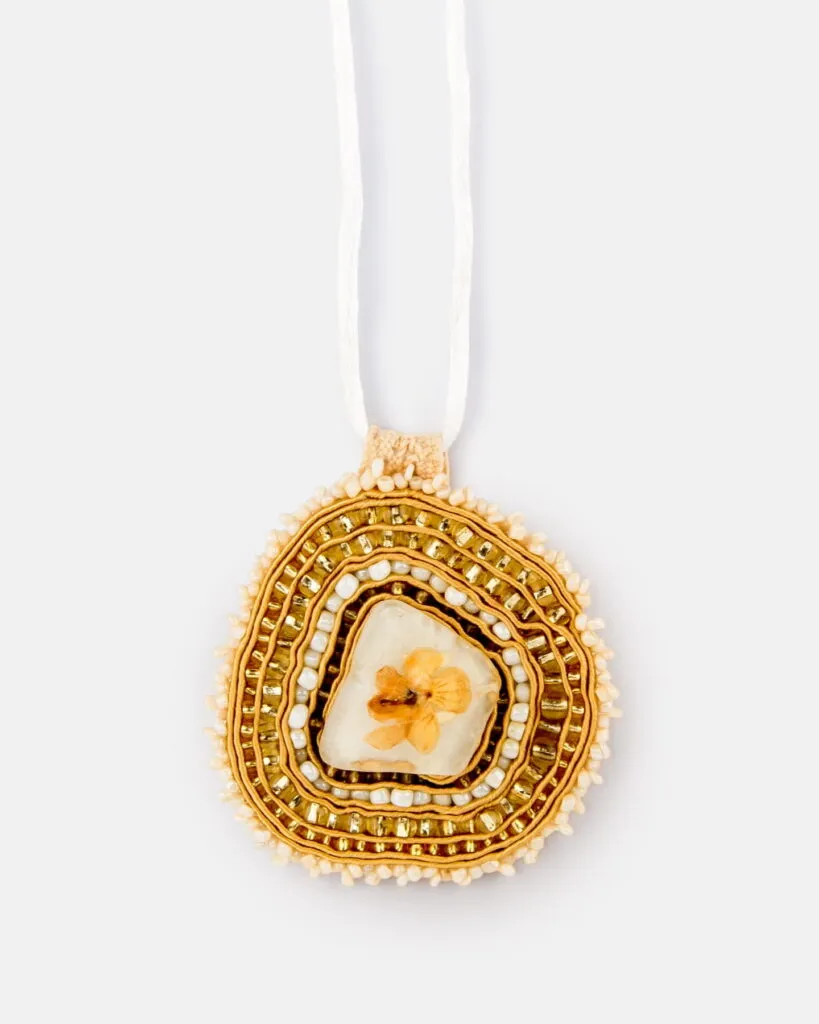 Wisiorek z koralików, w którym znajduje się półprzezroczysty kamień z wciśniętym kwiatem, otoczony koncentrycznymi warstwami złotych i kremowych koralików.